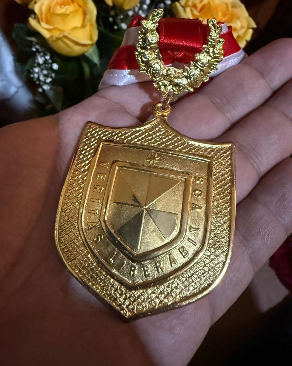 Medalla dorada de la Universidad de San Martín de Porres, con el escudo y el lema Veritas liberabit vos, sostenida en la mano.